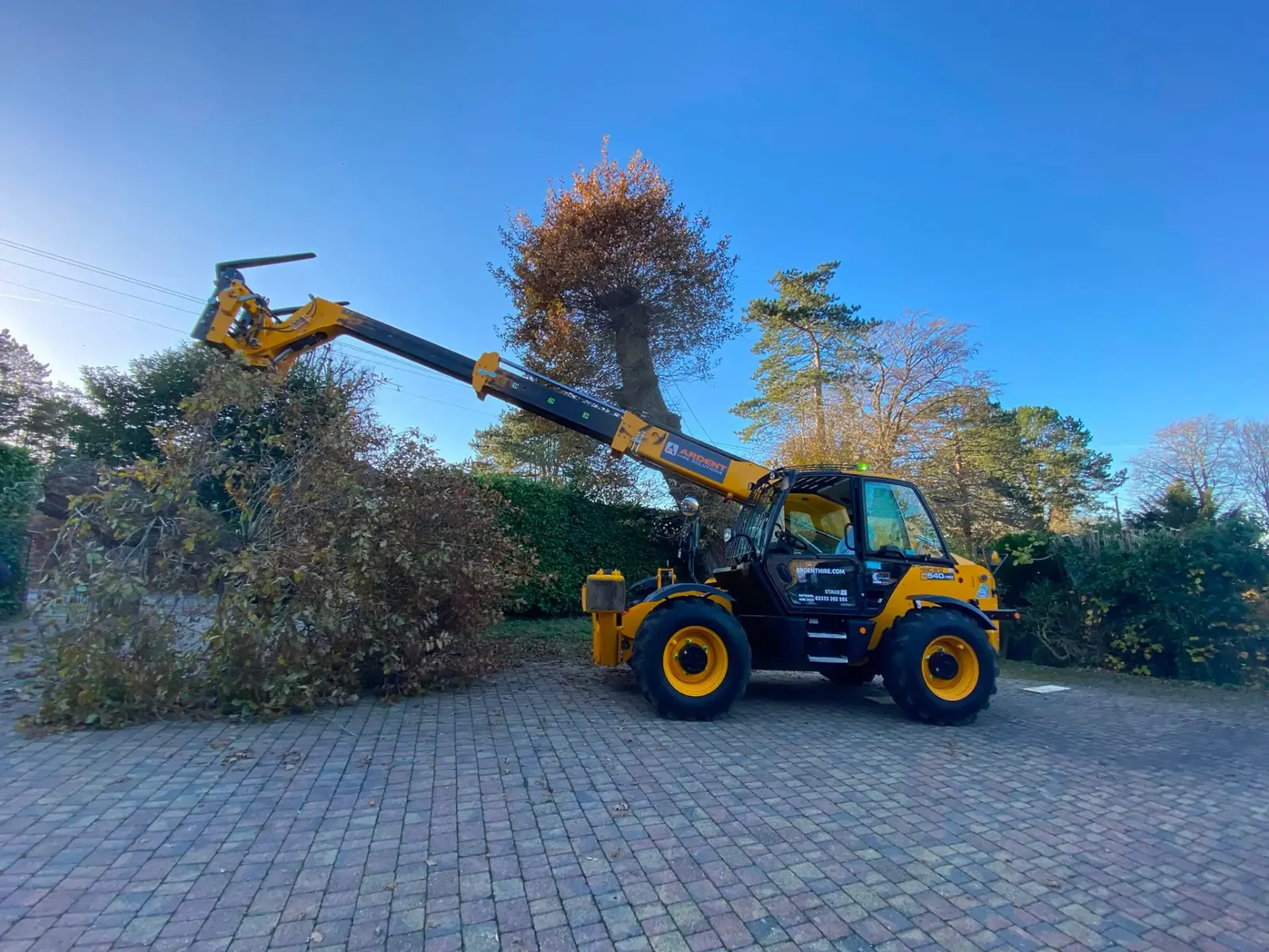 Telehandler lifting storm-damaged hedge and tree debris from a driveway